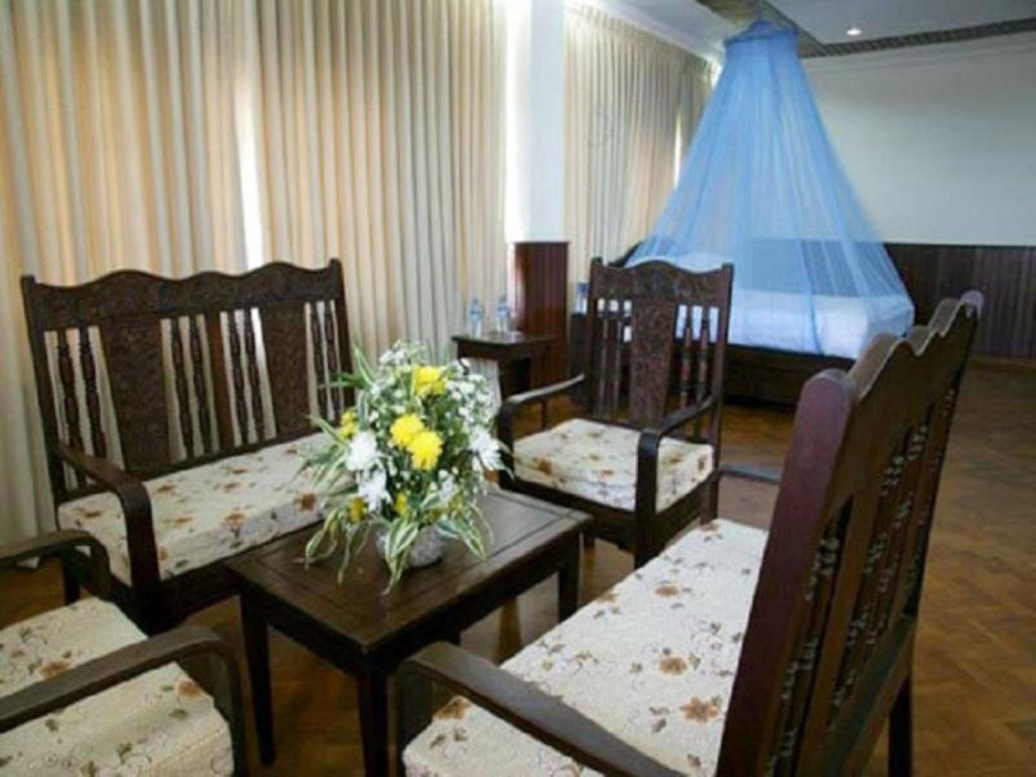 A room with four wooden chairs and a table, a floral centerpiece, and a mosquito net over a bed.