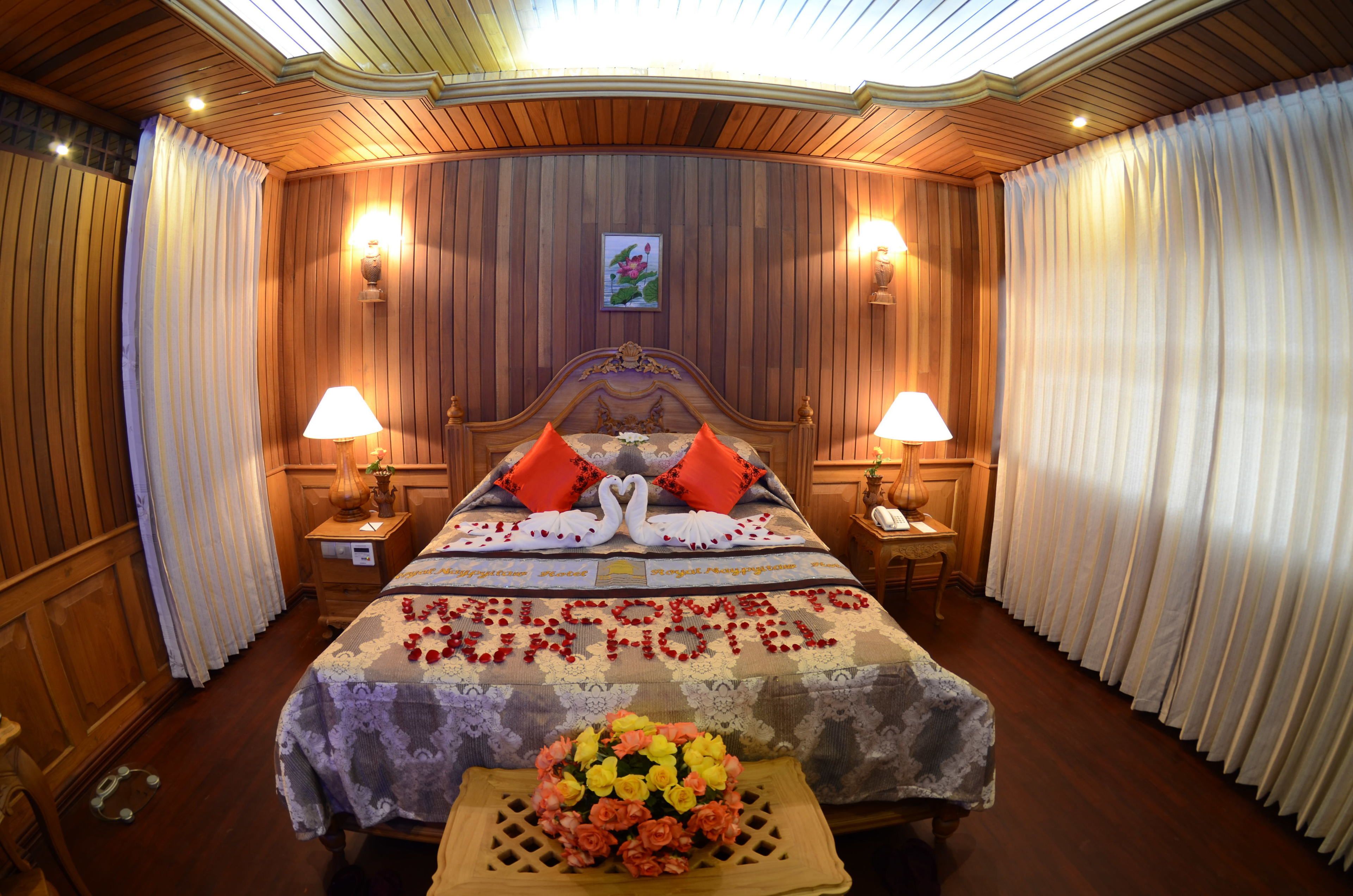 A hotel room with a wooden paneled wall, a bed with a floral bedspread, two bedside lamps, and a bouquet of flowers on a side table.