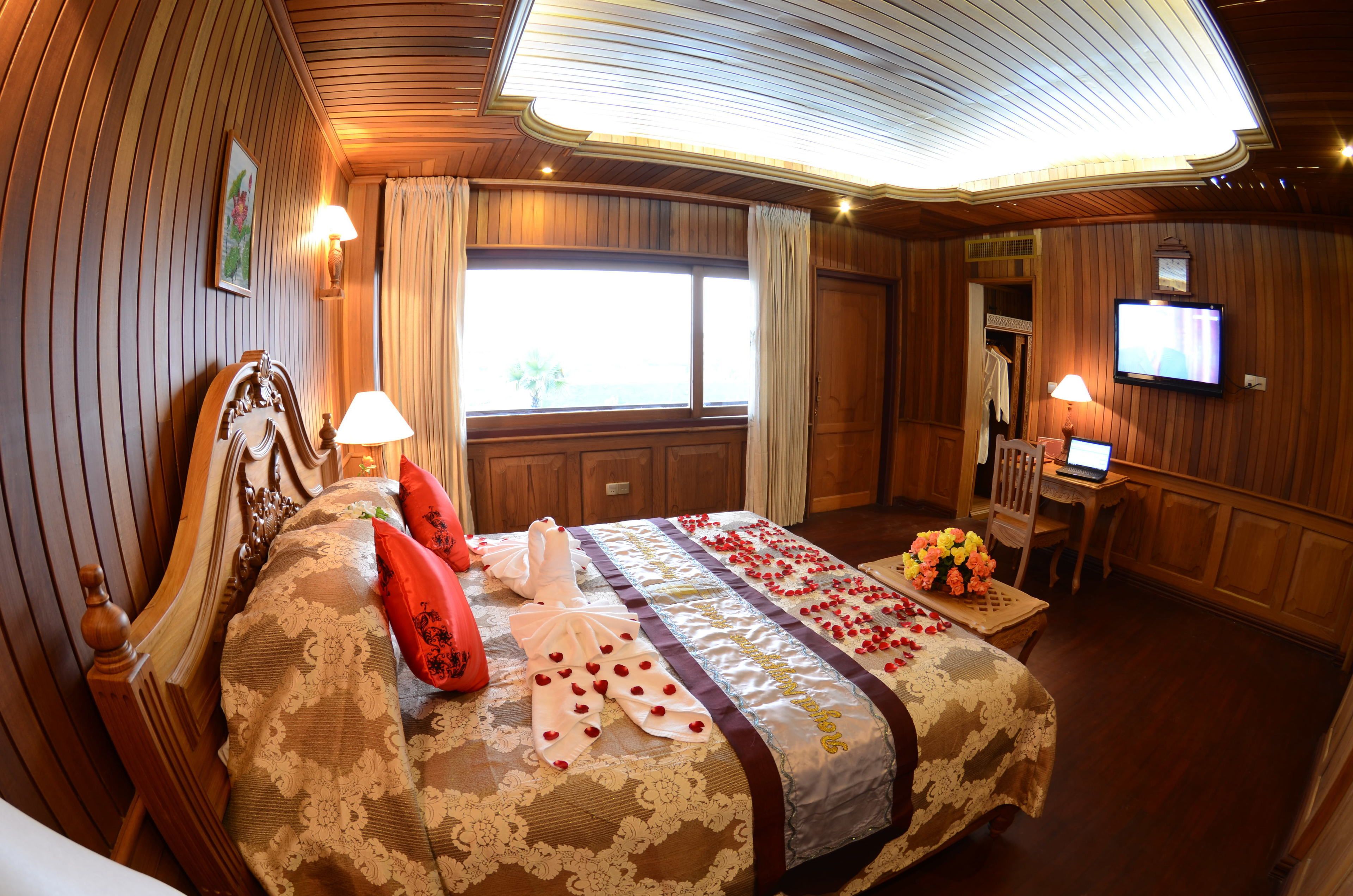 Een hotelkamer met een groot bed, een houten hoofdeinde, een bureau met een computer, een televisie aan de muur en een raam met gordijnen.