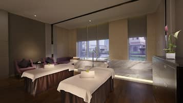 A modern spa room with two massage tables, a seating area, and a large window offering a city view.