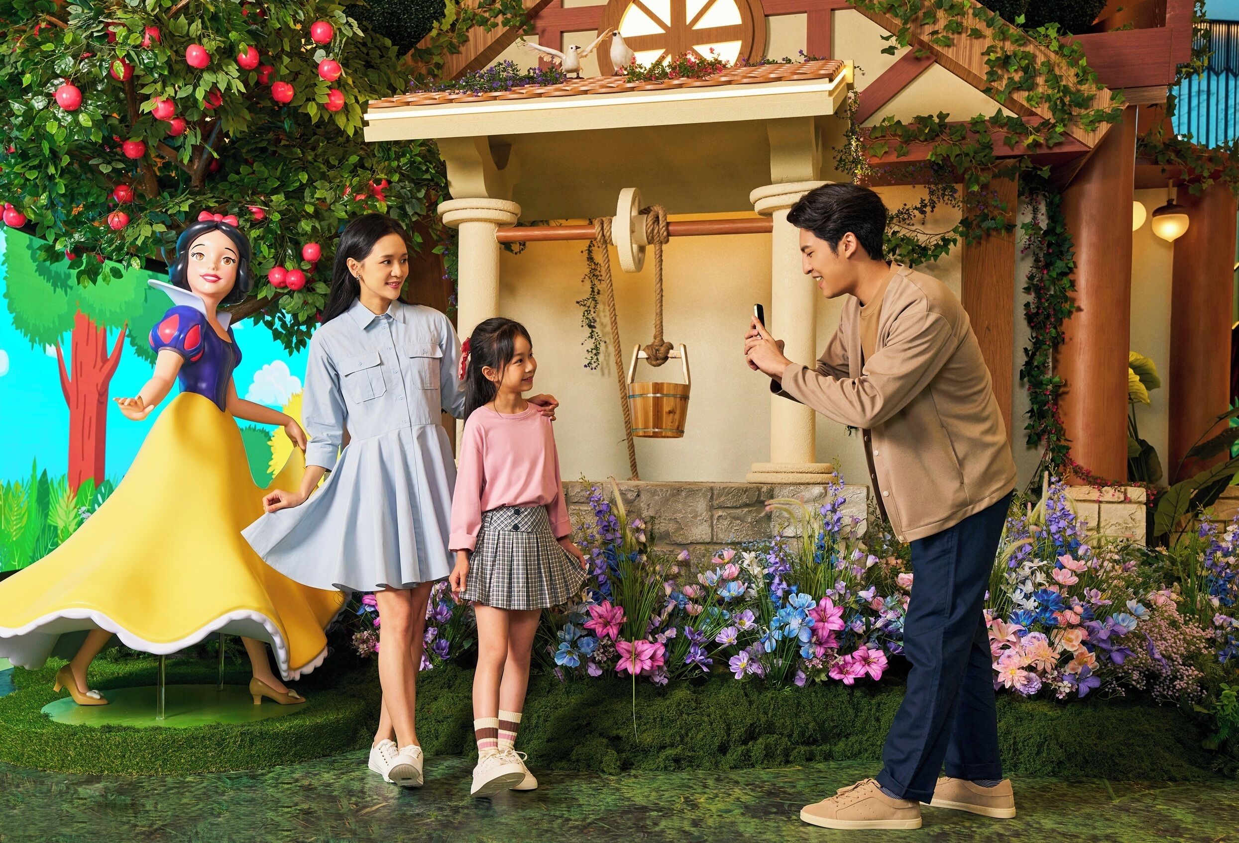 A family with a child and a woman in a blue dress standing in front of a building with a tree and flowers.
