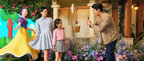 A family with a child and a woman in a blue dress standing in front of a building with a tree and flowers.