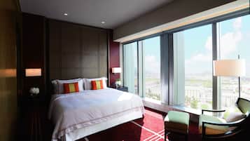 Una habitación de hotel con una cama grande, una zona de estar con una silla, una mesita de noche y vistas a la ciudad a través de amplias ventanas.