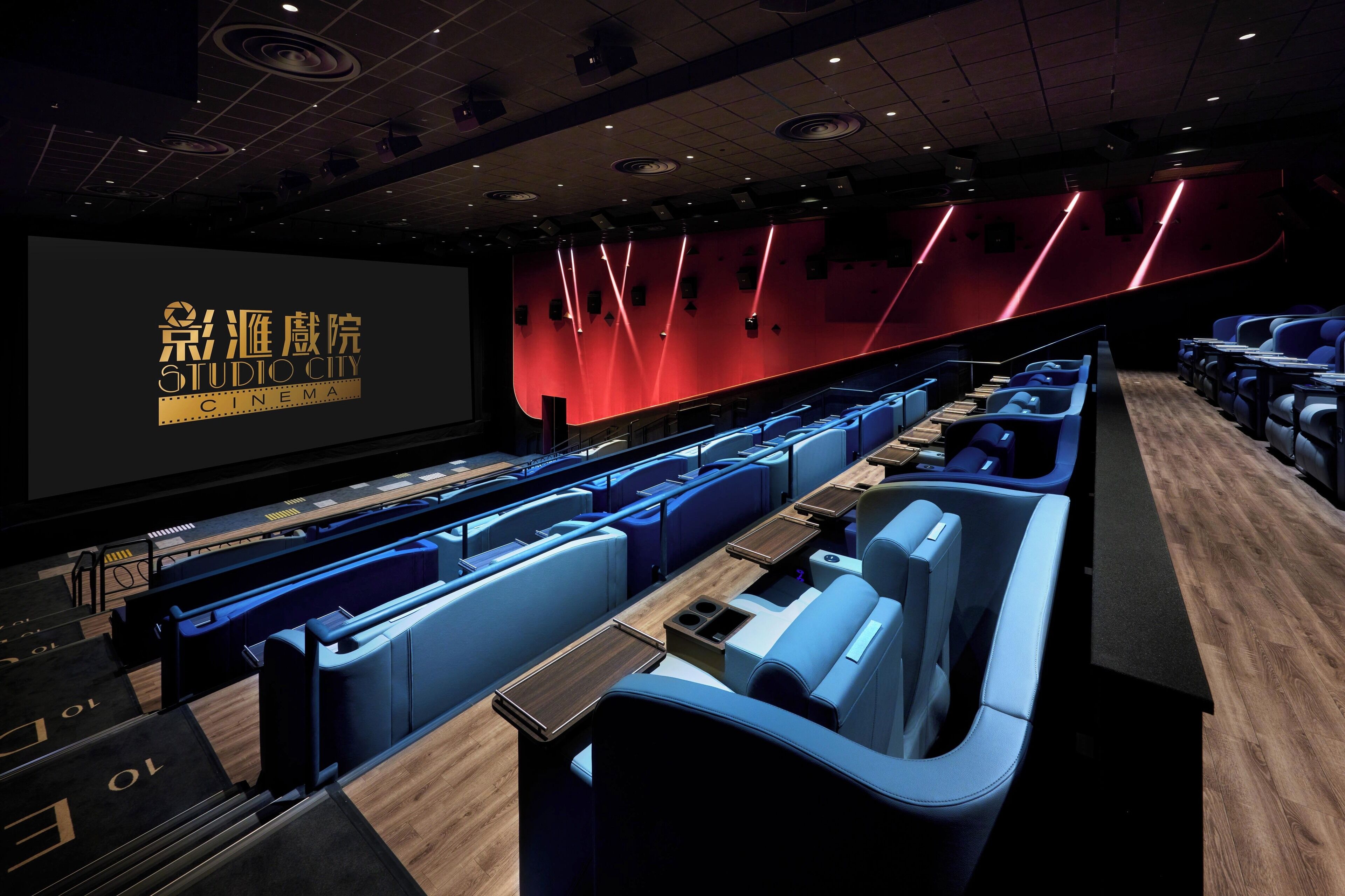 A cinema with rows of blue seats, a large screen, and a red wall with light fixtures.