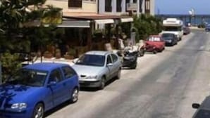 A coastal street with multi-story buildings, parked cars, and a clear view of the sea. - Evagellina (Rethymno)
