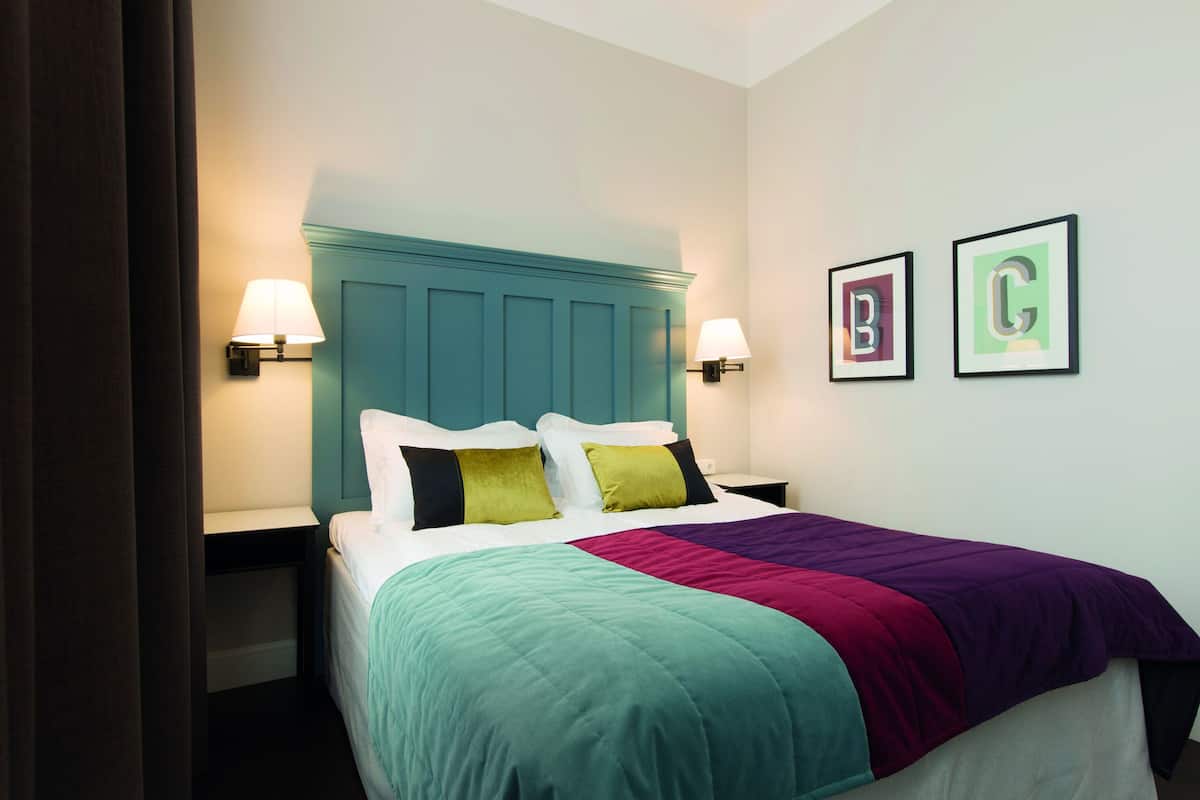 Cama bem arrumada com cabeceira verde-água, duas luminárias de cabeceira e um edredom colorido.