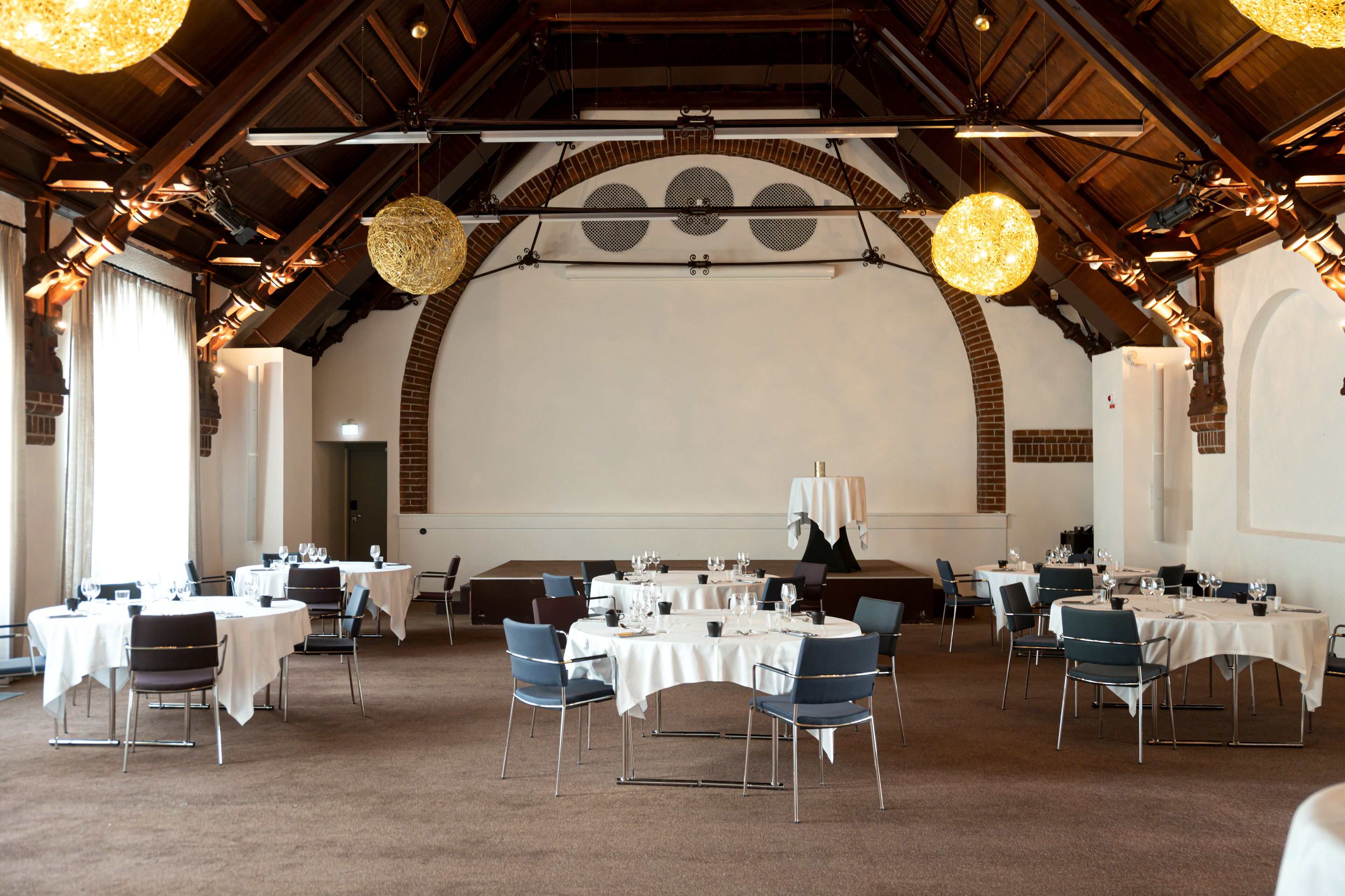 A spacious dining area with multiple tables and chairs, high ceilings with exposed beams, and hanging spherical light fixtures.