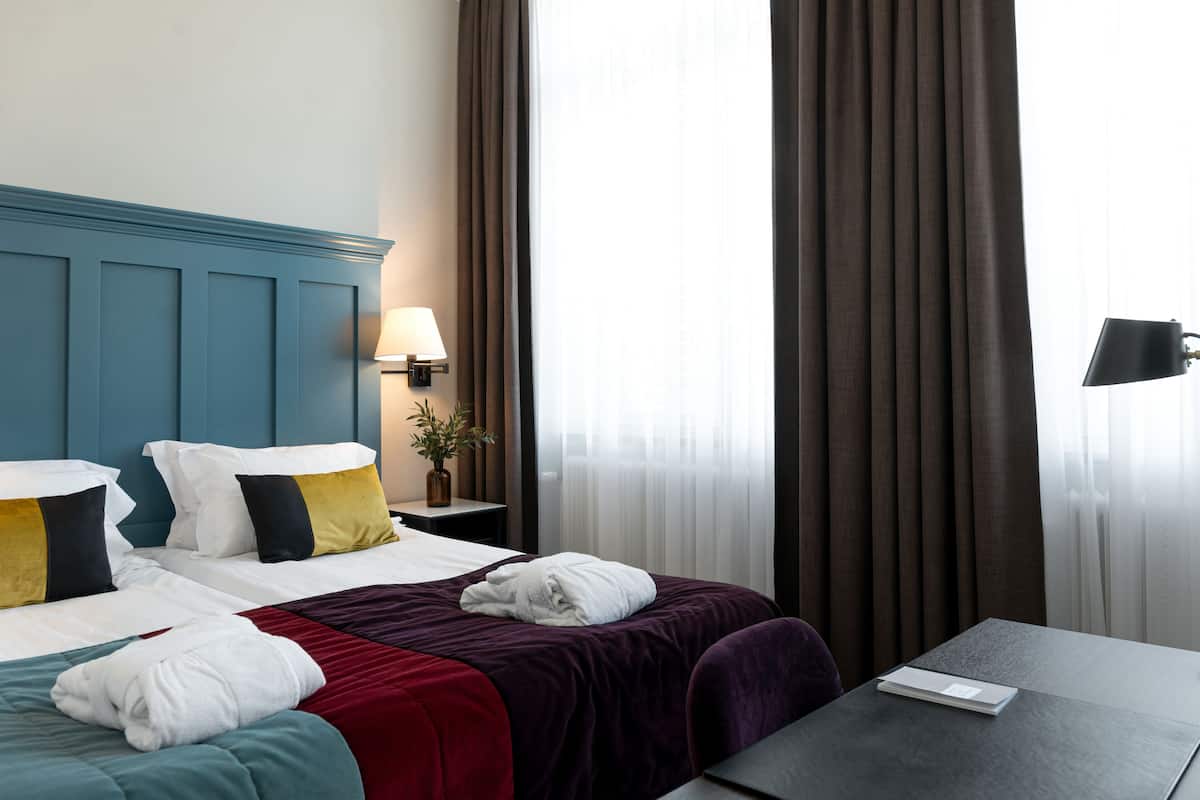 Quarto de hotel com duas camas, uma mesa de cabeceira com abajur, uma mesa e vista para o exterior através de cortinas.