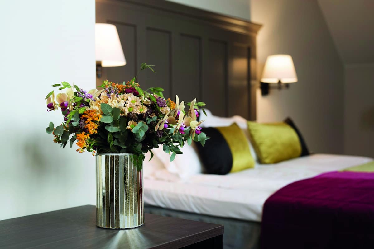 Quarto de hotel com uma cama, mesa de cabeceira com um vaso de flores e duas luminárias.