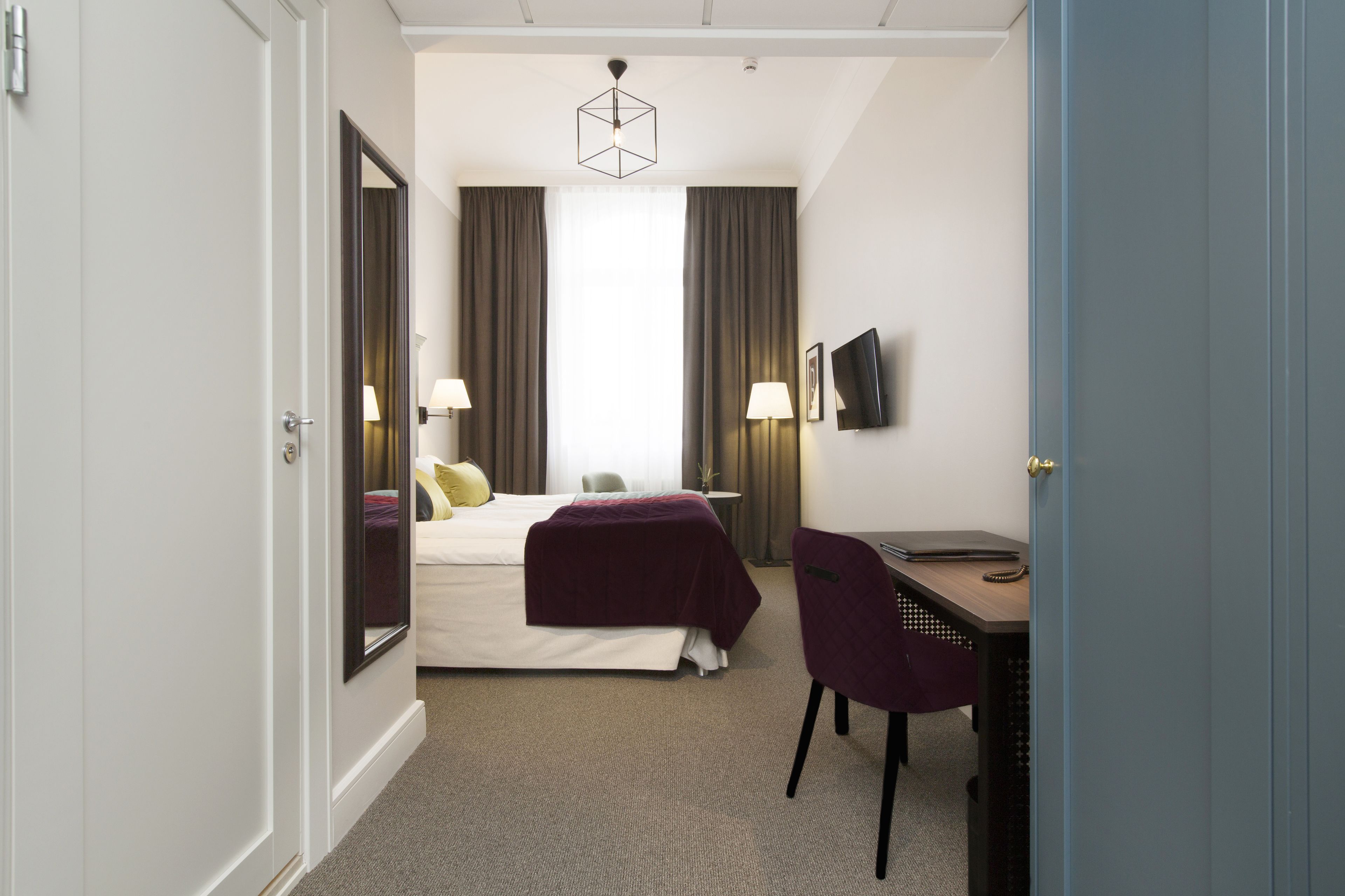 Une chambre d’hôtel avec un lit, un bureau, une chaise, une télévision et une fenêtre avec des rideaux.