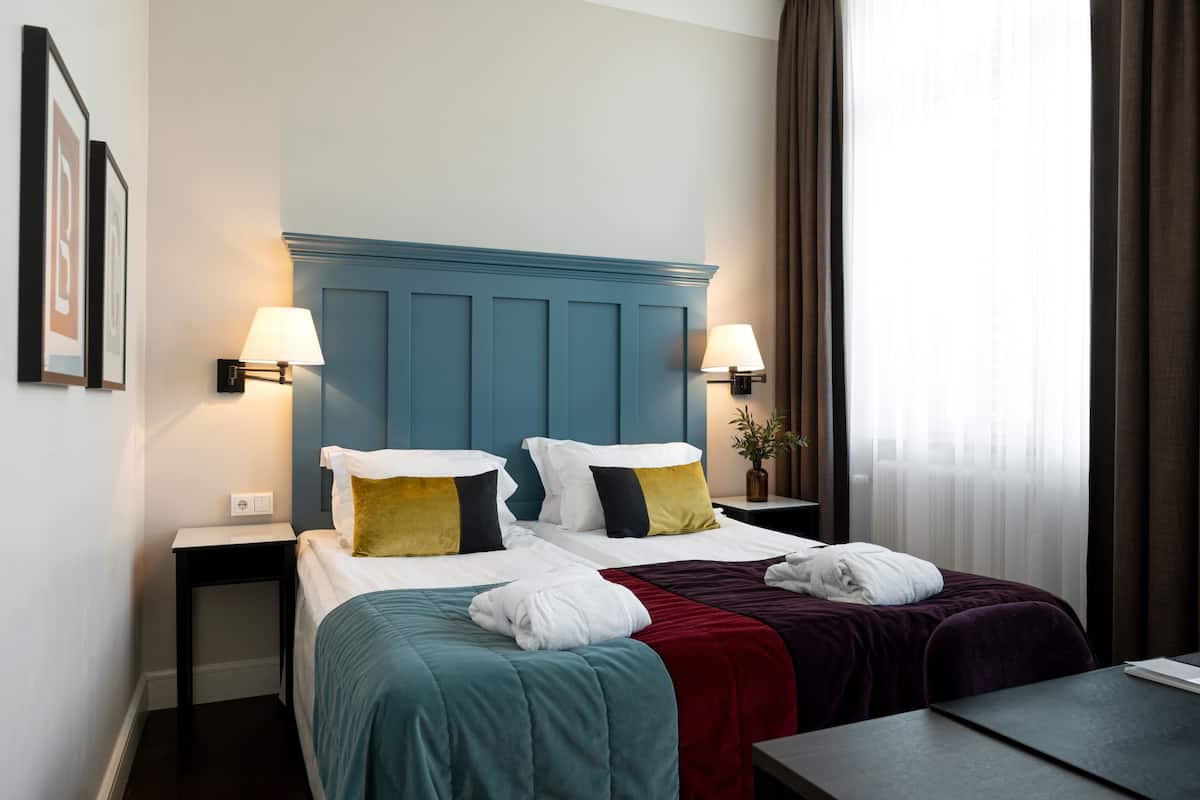 Quarto de hotel com duas camas, cabeceira azul, abajures, uma mesa pequena e uma janela com cortinas.