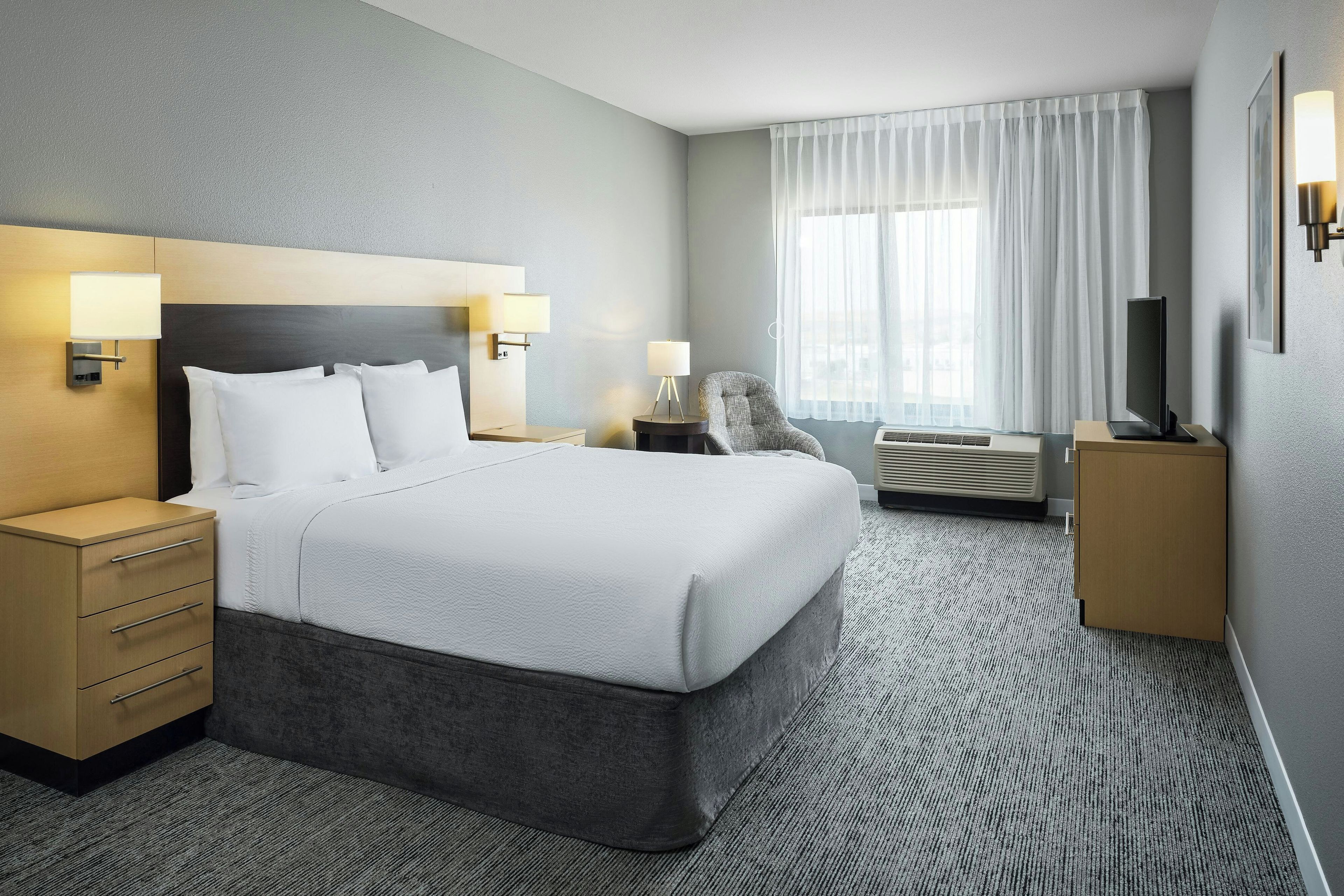 A hotel room with a large bed, bedside tables, a chair, a desk, a flat-screen TV, and a window with curtains.