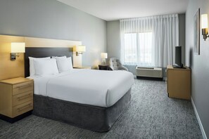 A hotel room with a large bed, bedside tables, a chair, a desk, a flat-screen TV, and a window with curtains.