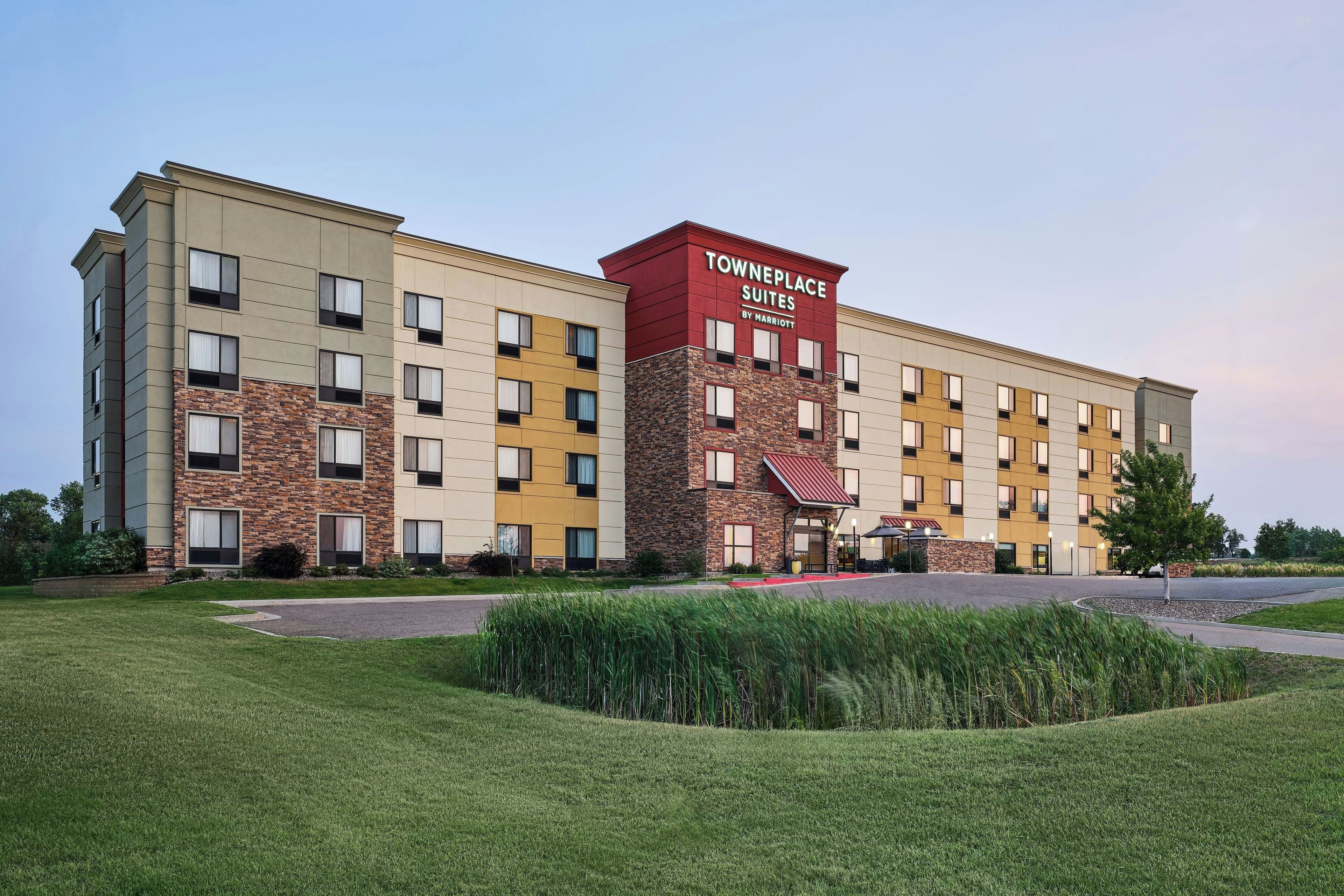 A multi-story hotel with a prominent red and beige facade, a landscaped front yard, and a parking area.