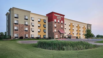 A multi-story hotel with a prominent red and beige facade, a landscaped front yard, and a parking area.