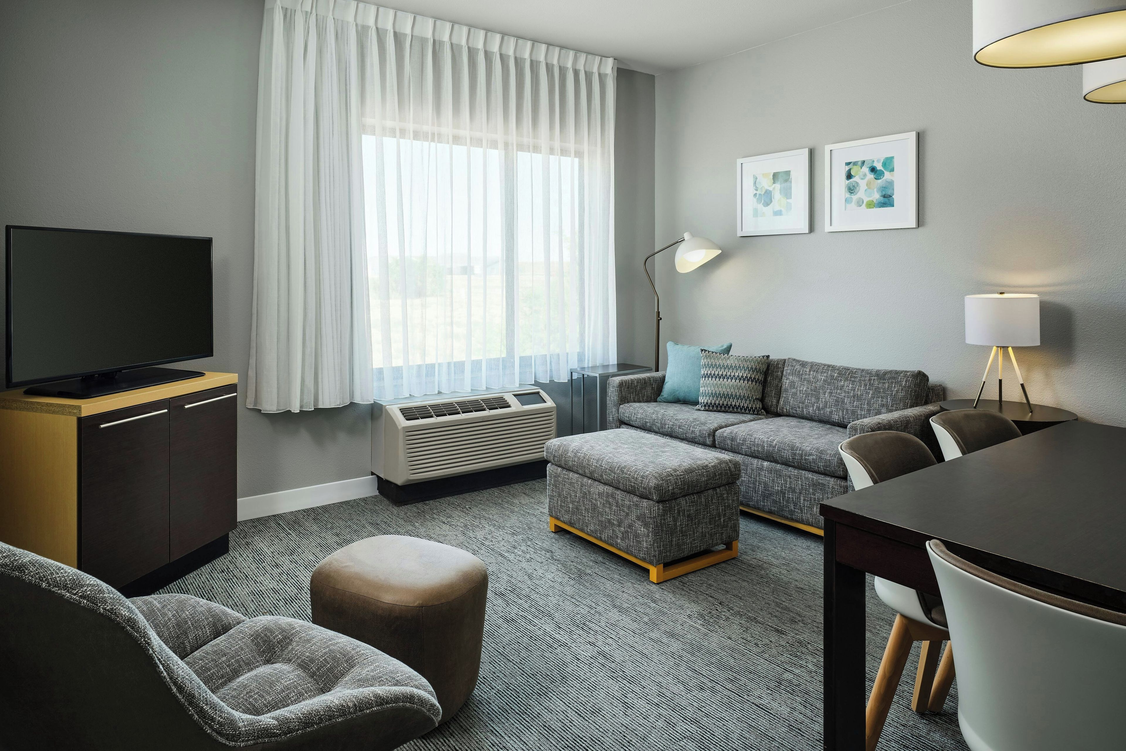 Ein modernes Hotelzimmer mit einem Flachbildfernseher, einer Sitzecke, einem Esstisch und einem großen Fenster mit leichtem Vorhang.