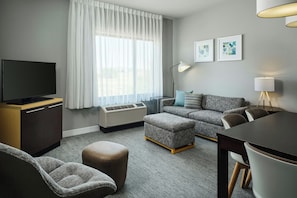 Una habitación de hotel moderna con una televisión de pantalla plana, un sofá, una mesa de comedor y un ventanal con cortinas translúcidas.