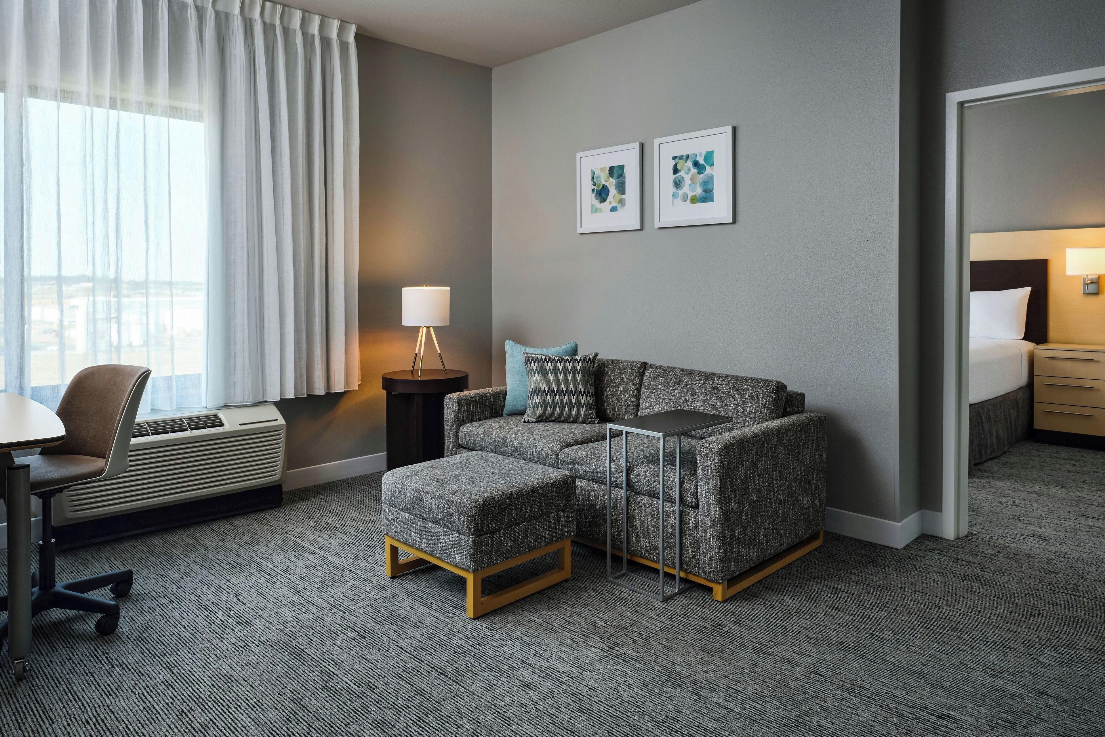 Une chambre d’hôtel équipée d’un canapé, d’un pouf, d’un bureau, d’une chaise, d’une lampe et d’une œuvre d’art accrochée au mur.