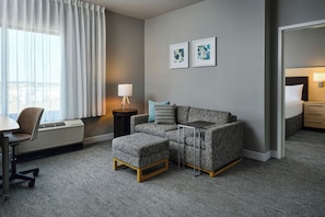 A hotel room with a sofa, ottoman, desk, chair, lamp, and artwork on the wall.