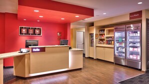 Uma área de recepção de hotel com parede vermelha, balcão de recepção, computador e frigorífico abastecido com bebidas.