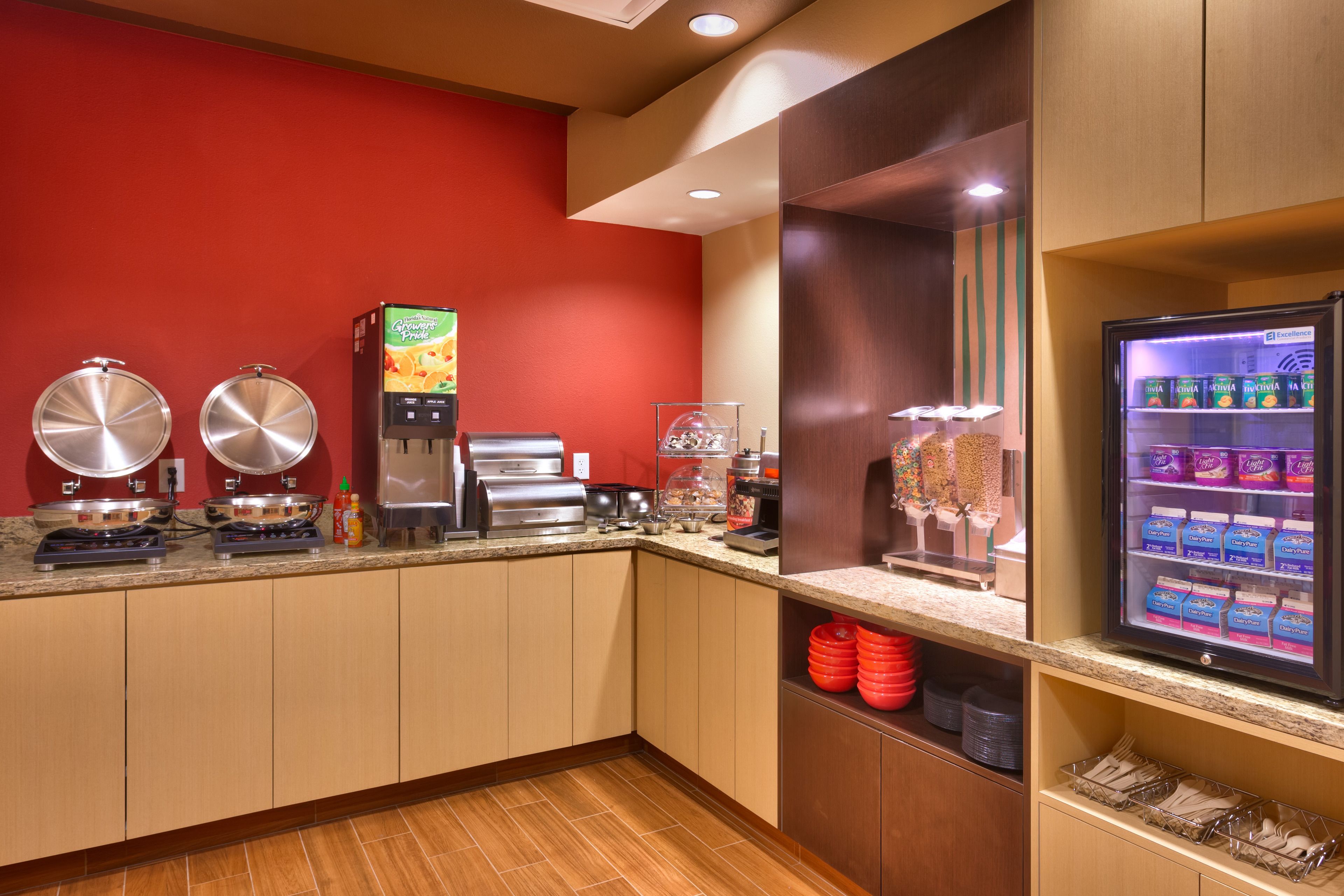 A self-service breakfast area with coffee machines, a juice dispenser, and a refrigerated display of dairy products.