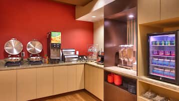 A self-service breakfast area with coffee machines, a juice dispenser, and a refrigerated display of dairy products.