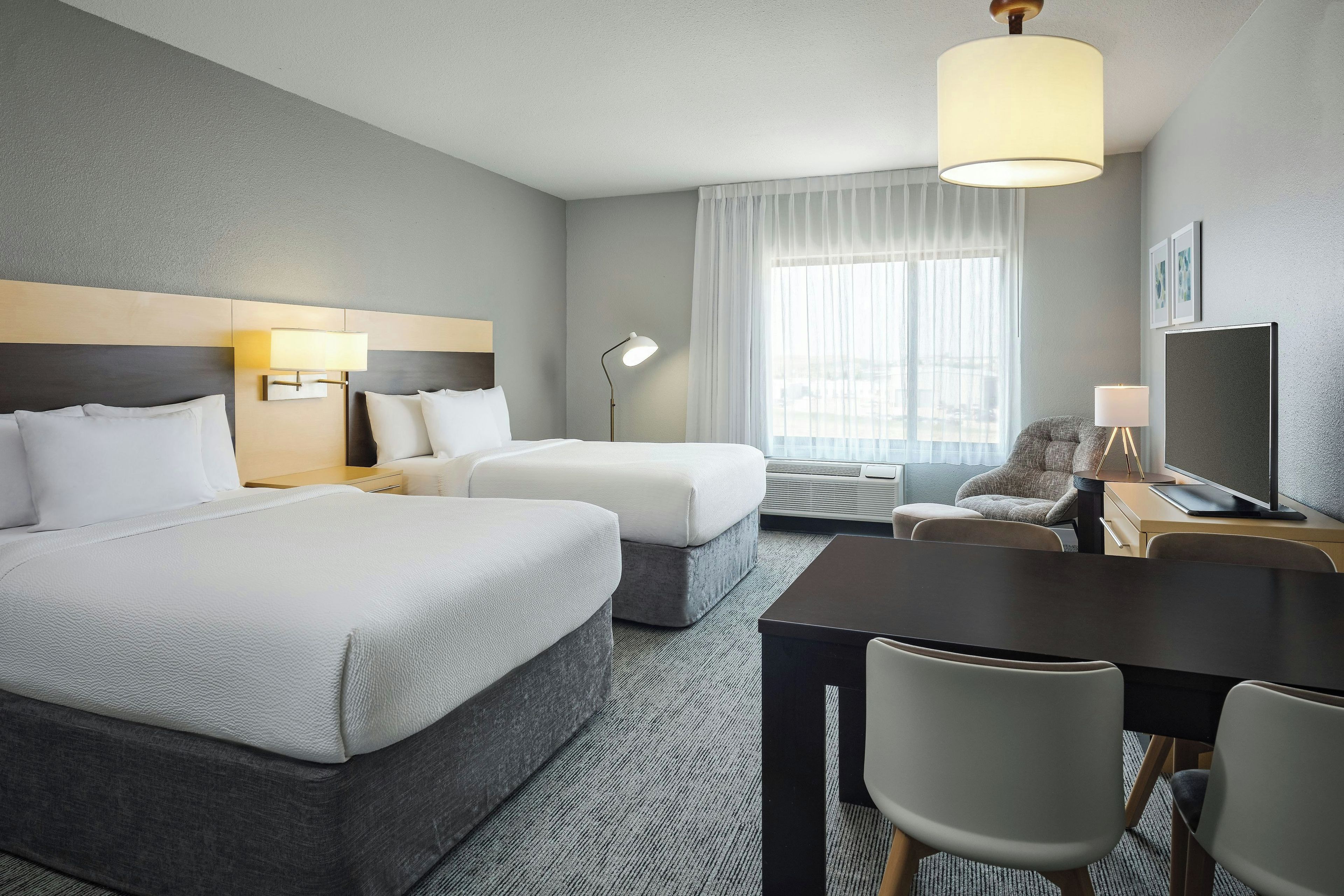 A hotel room with two beds, a desk with a chair, a lamp, and a large window with sheer curtains.