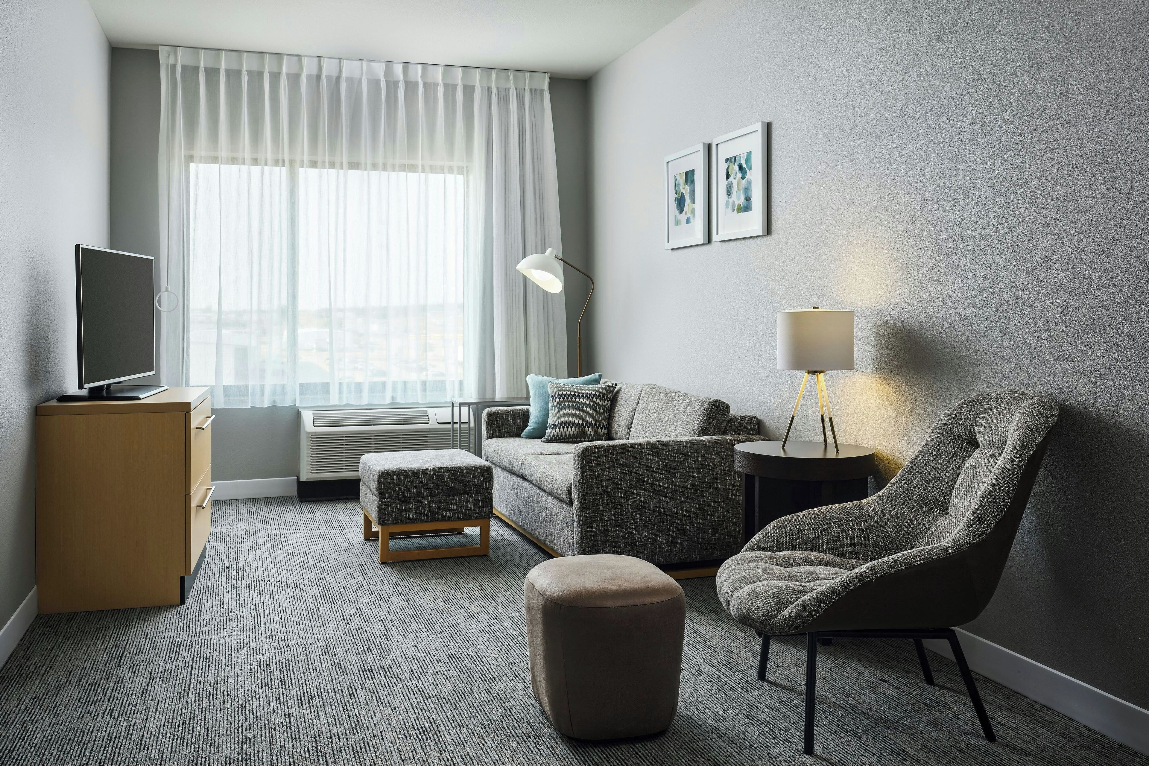 A modern hotel room with a flat-screen TV, a sofa, an ottoman, a chair, a lamp, and a large window with sheer curtains.