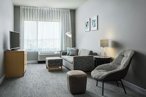 Een moderne hotelkamer met een flatscreen televisie, een bank, een poef, een stoel, een lamp en een groot raam met dunne gordijnen.