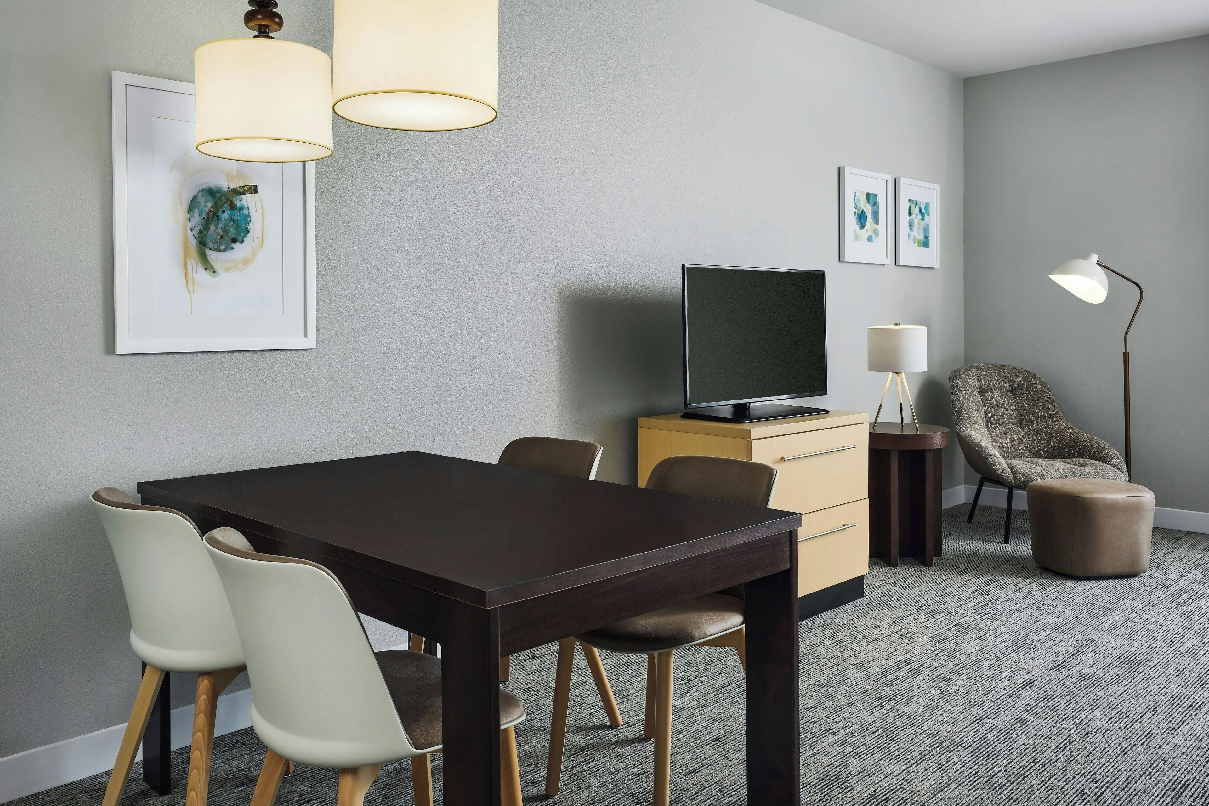 A modern hotel room with a dark wooden dining table, white chairs, a flat-screen TV, a small cabinet, a floor lamp, and artwork on the walls.