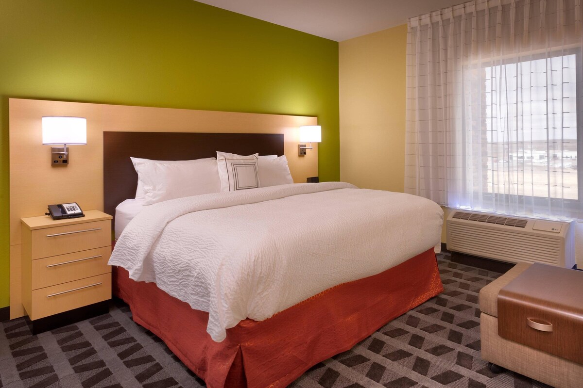 A hotel room with a large bed, two bedside lamps, a telephone, a heater, and a window with blinds.