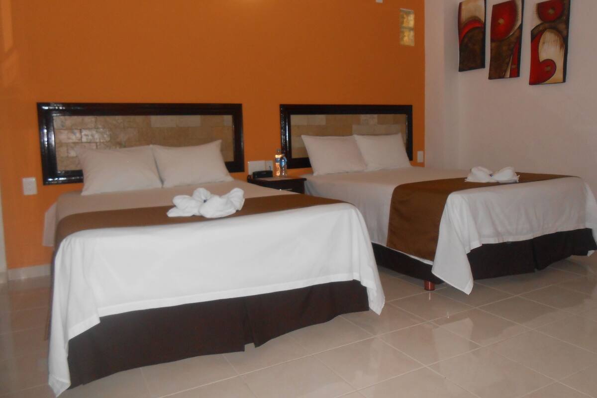 A hotel room with two beds, each with a headboard, white bedding, and a brown skirt. There are two nightstands with lamps, a water bottle on one, and a red cup on the floor. The walls are orange, and there are three framed artworks on the wall.