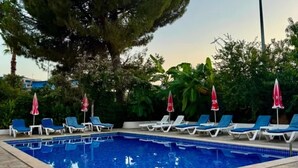 Una piscina con sillones de descanso y sombrillas, rodeada de árboles y un cielo despejado.