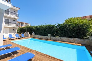 A swimming pool with blue lounge chairs, a stone wall, and a building with balconies. - Karras Livin (Zakynthos)