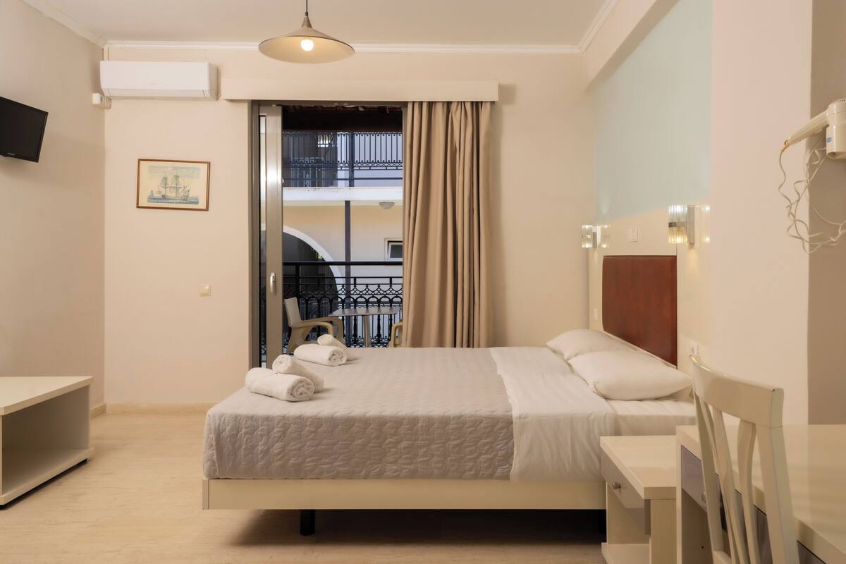 A hotel room with a bed, a desk, a chair, a TV, and a balcony with a railing.