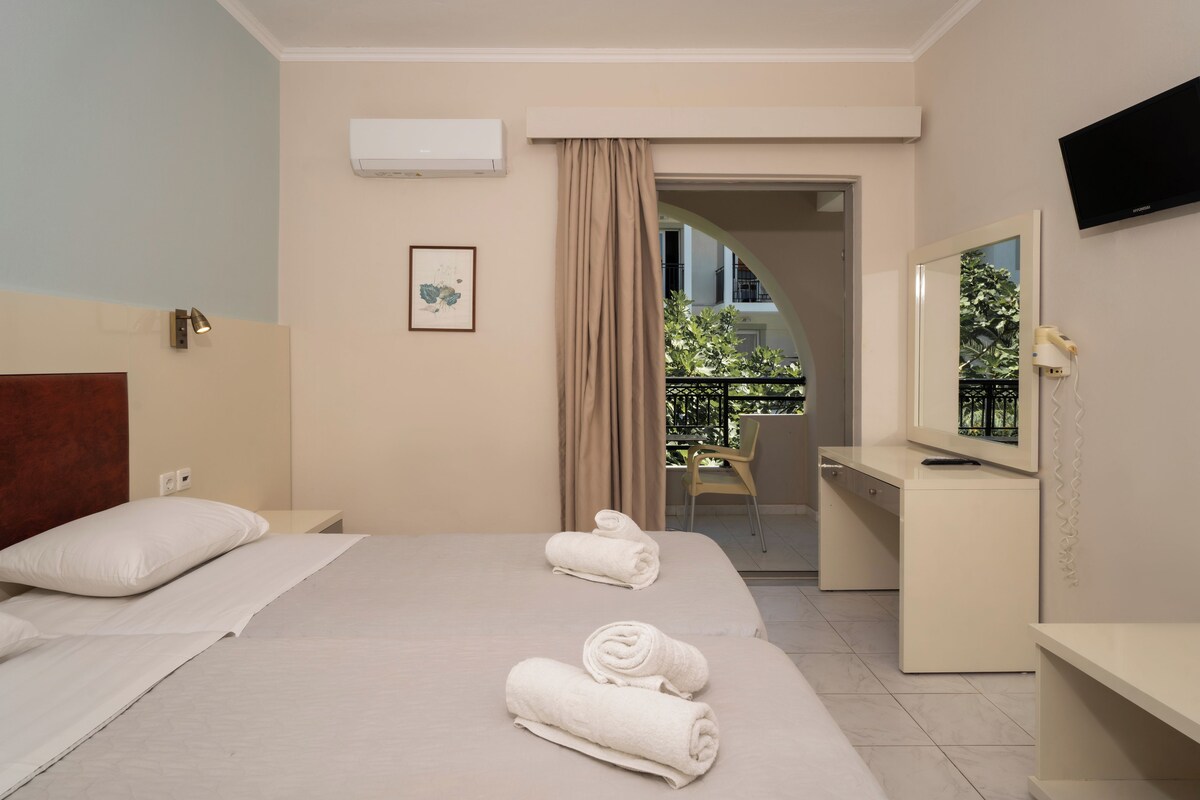 Quarto de hotel com cama, escrivaninha, cadeira, televisão e varanda com plantas.