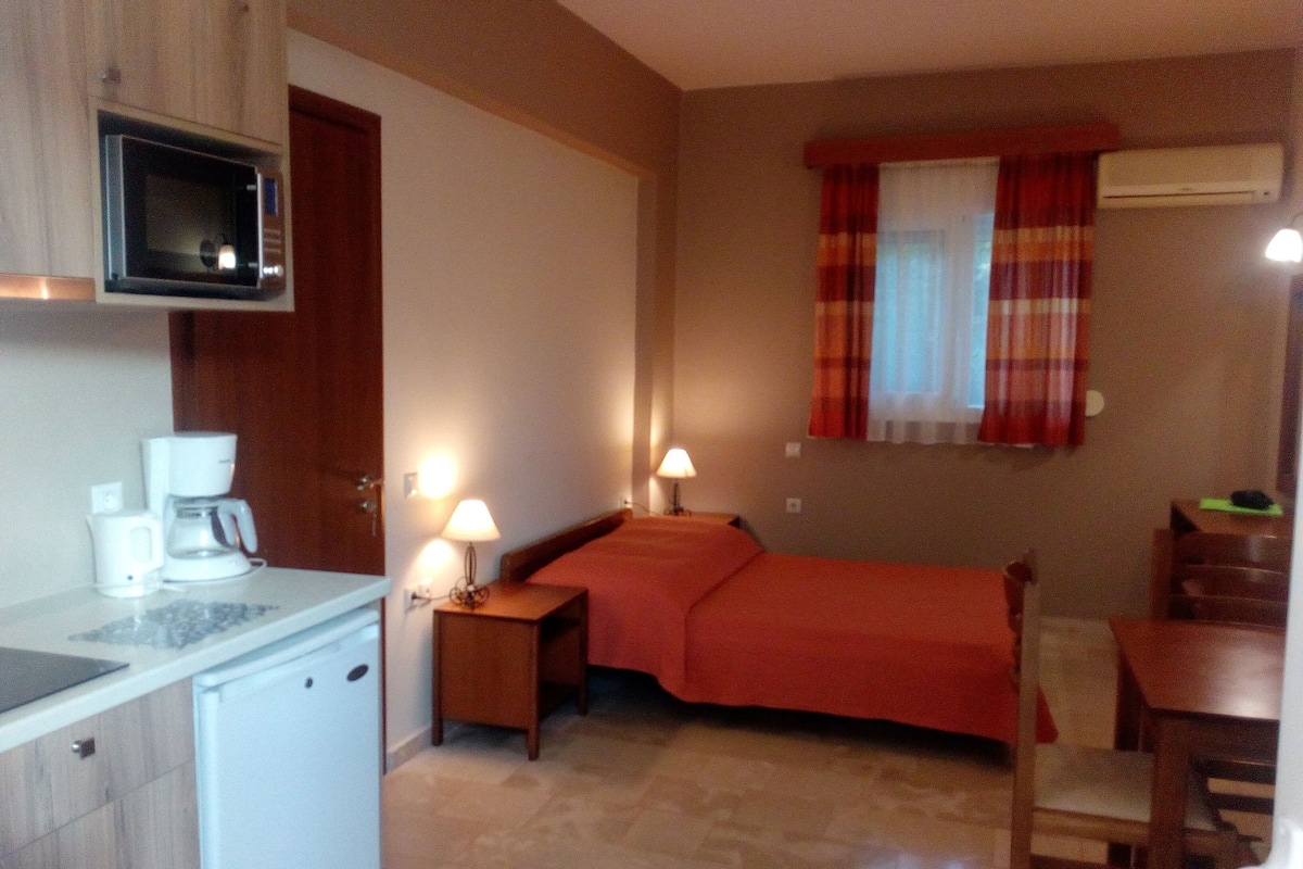 A compact hotel room with a kitchenette, a bed with a red bedspread, a small table, and a chair.