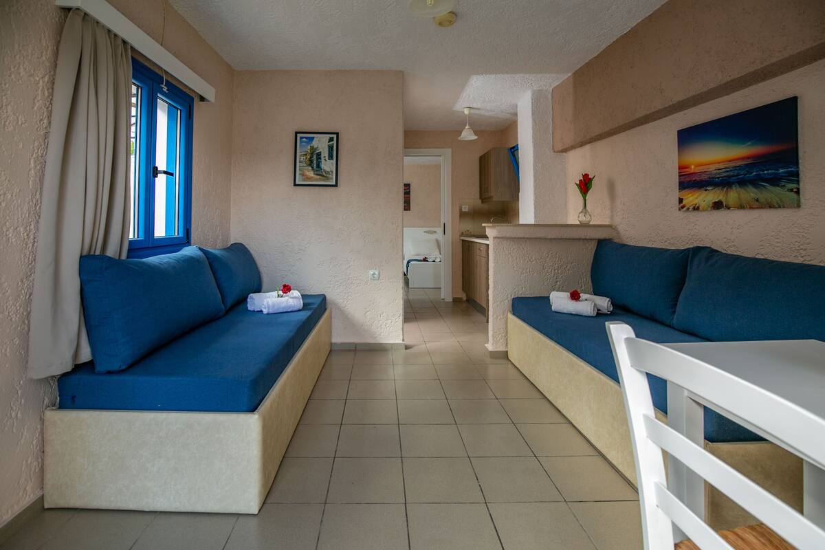 Ein Zimmer mit zwei blauen Sofas, einem weißen Tisch und einer Küchenzeile im Hintergrund.