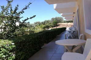 A patio with white plastic chairs and tables, surrounded by a hedge and greenery, with a clear view of the landscape in the background.