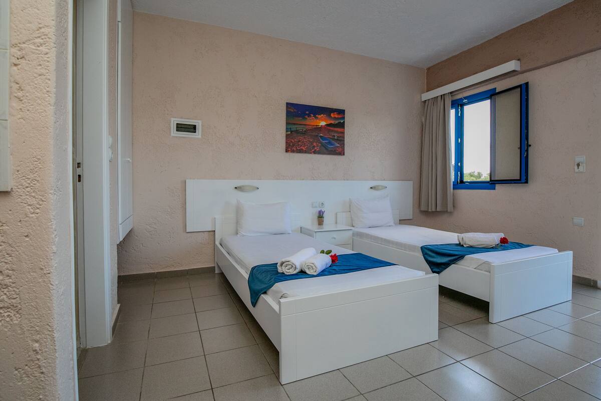 Ein Hotelzimmer mit zwei Betten, einem Bild an der Wand, einem Fenster mit Vorhängen und einem gefliesten Boden.