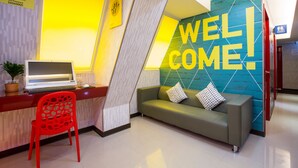 Un área de recepción con una laptop sobre un escritorio, una silla roja, un sofá verde con cojines y un mural que dice 'WELCOME'.