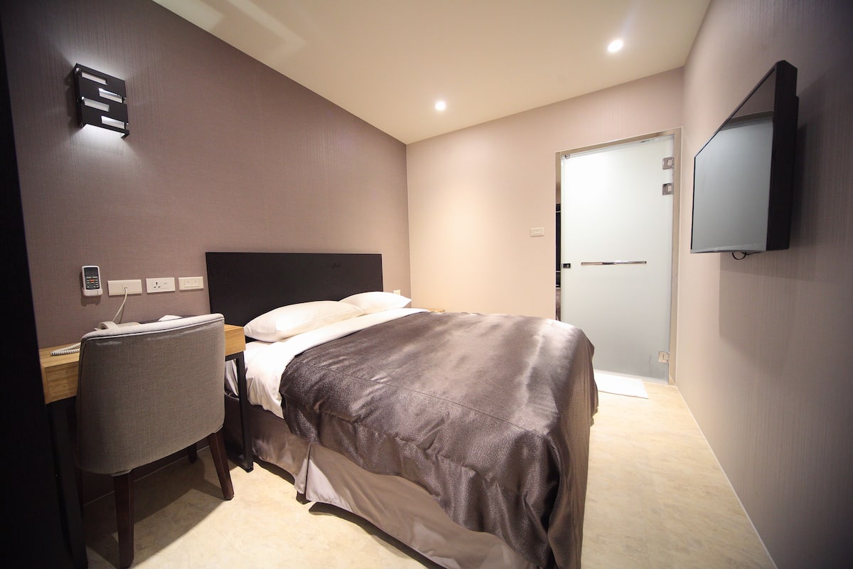A hotel room with a bed, a desk, a chair, a TV mounted on the wall, and a door.