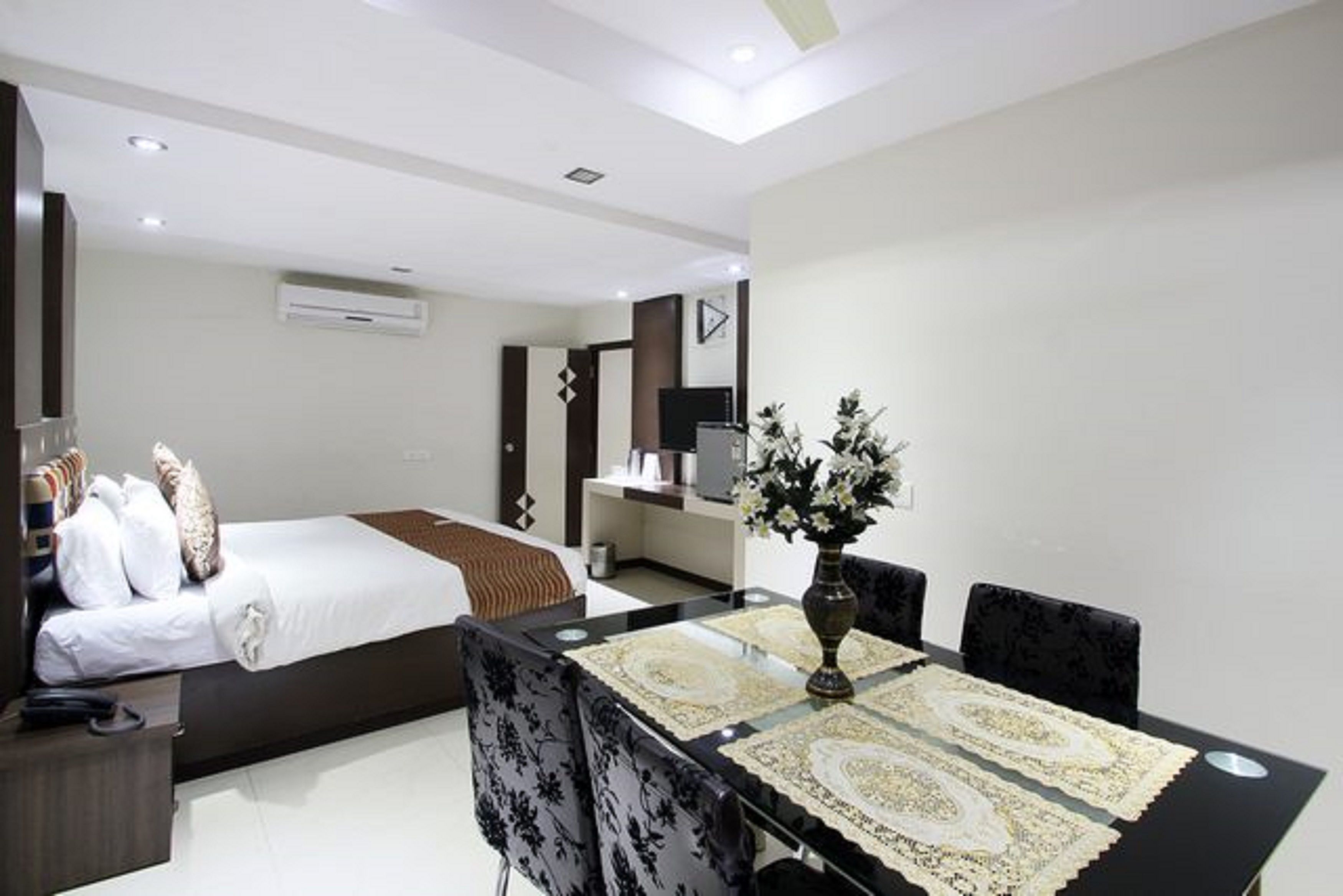A modern hotel room with a bed, a dining table with chairs, a TV, and a small kitchen area.