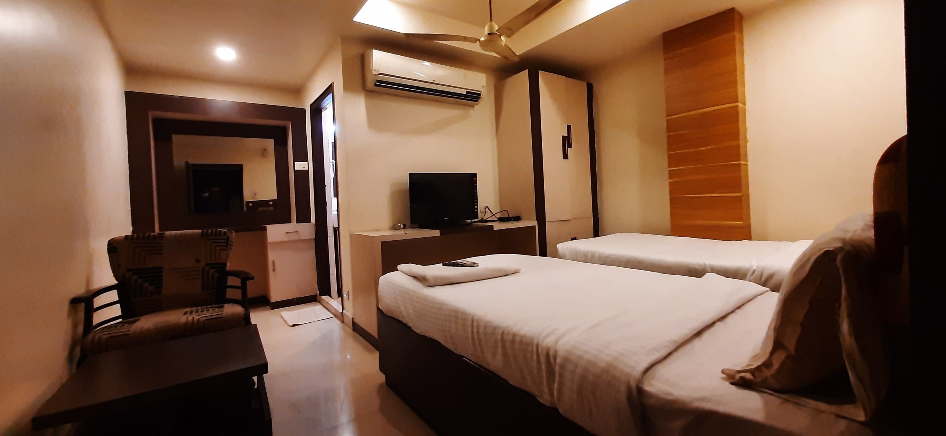 A hotel room with two beds, a desk, a chair, a TV, and an air conditioning unit.