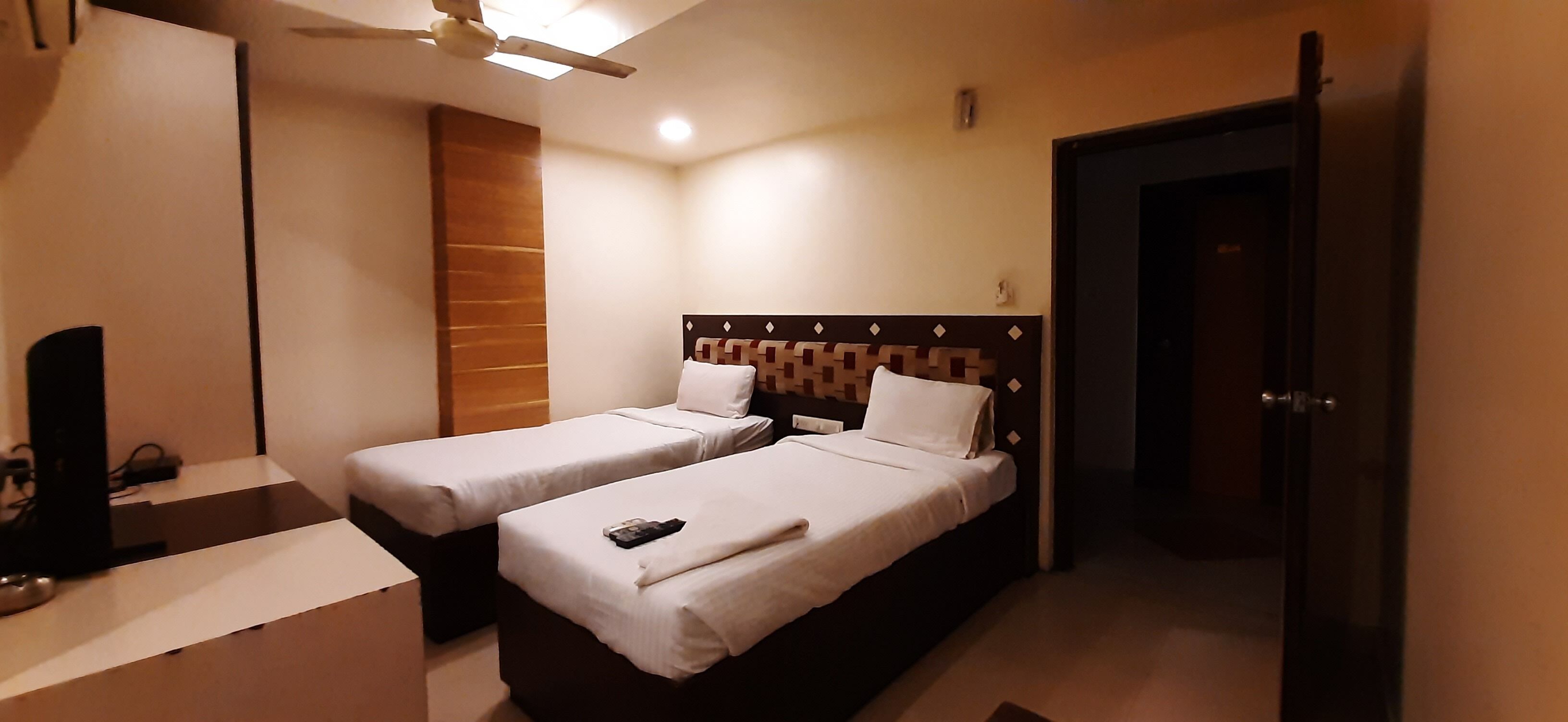 Habitación de hotel con dos camas individuales, un escritorio con silla, ventilador de techo y una puerta.