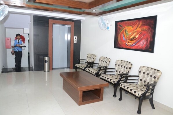 A reception area with a wooden coffee table, four patterned chairs, a painting on the wall, and a person standing near a door.