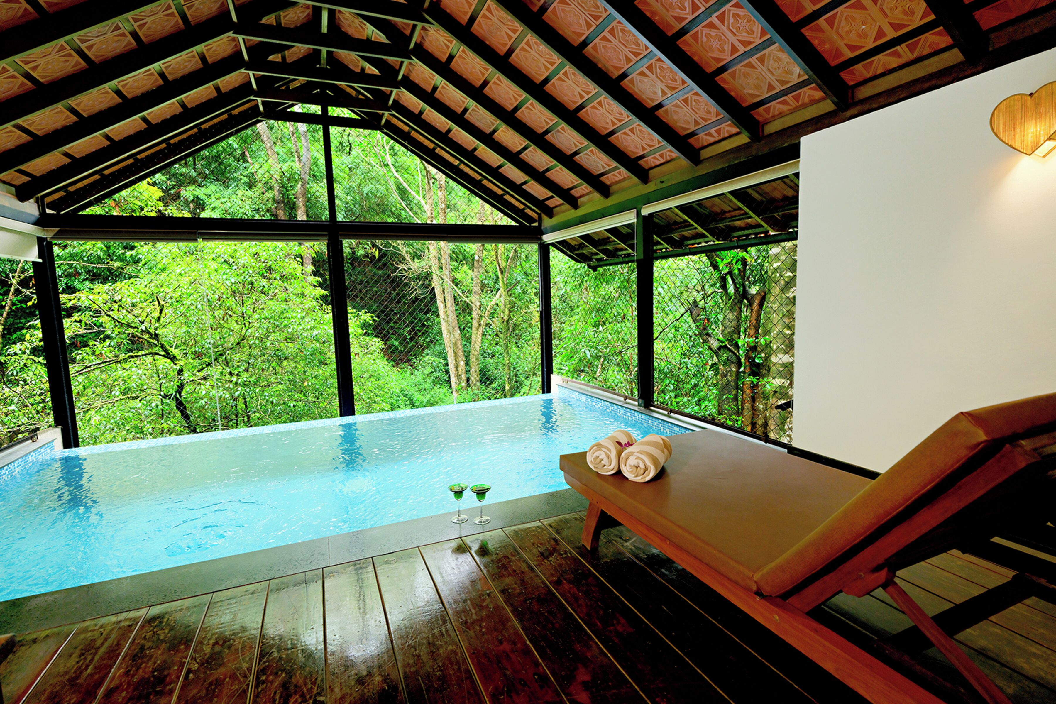 An infinity pool with wooden deck, a lounge chair, and a view of lush greenery.