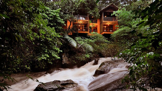 A multi-story building with balconies, surrounded by dense greenery and a flowing river. - Vythiri Resort (Vythiri)