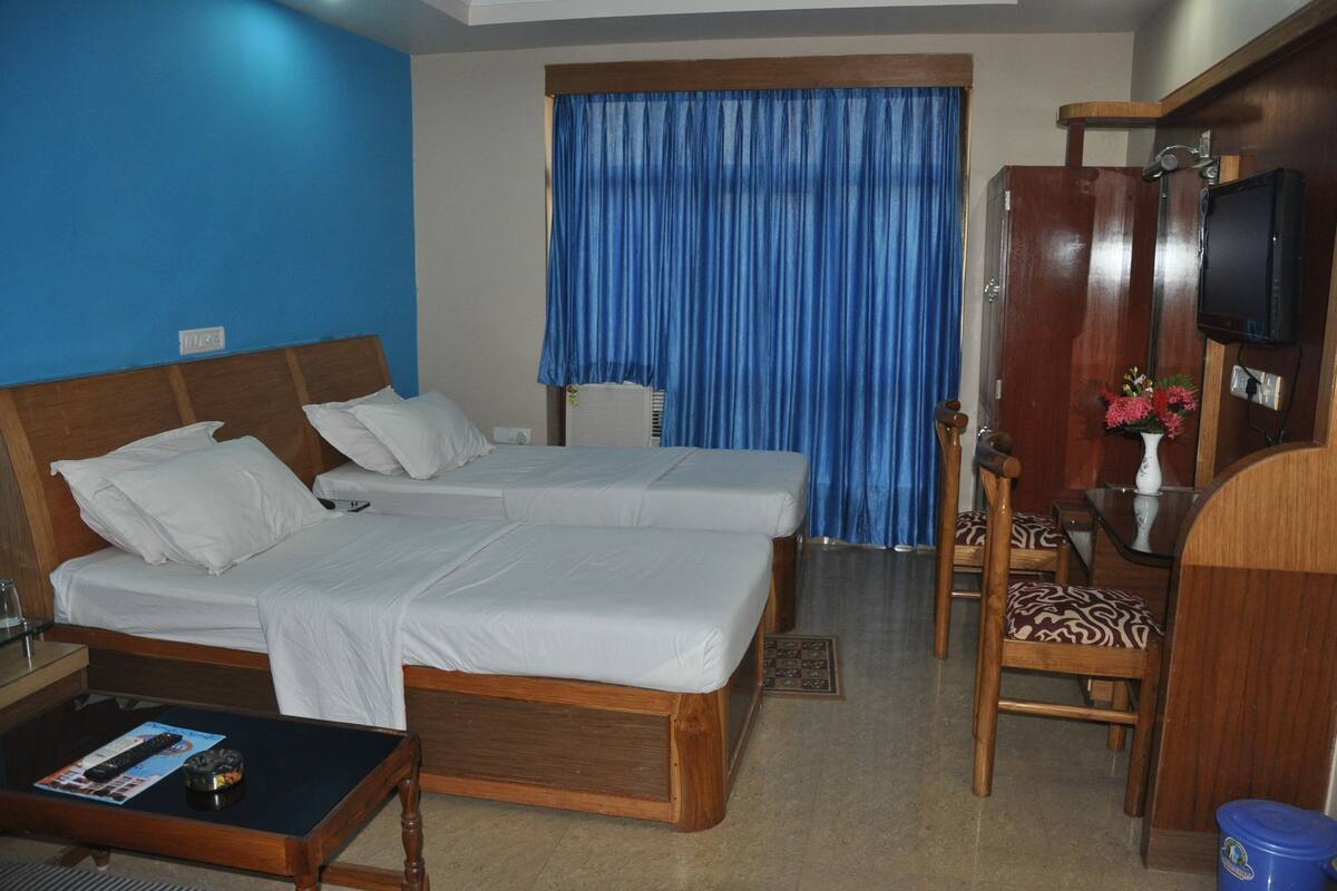 Habitación de hotel con dos camas, un escritorio de madera, una silla, un televisor y una ventana con cortinas azules.