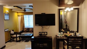 A hotel room with a flat-screen TV mounted on the wall, a dining table with chairs, a sofa, and a small table with a mirror and decorative items. - Hotel Pushpak (Bhubaneshwar)